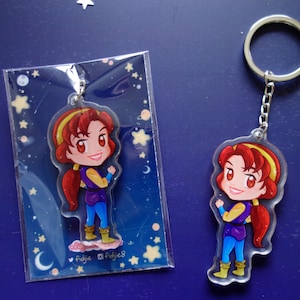 May include: A clear acrylic keychain featuring a cartoon character with red hair, a yellow headband, and a purple and blue outfit. The keychain is attached to a silver chain and ring. The character is also shown in a plastic sleeve.
