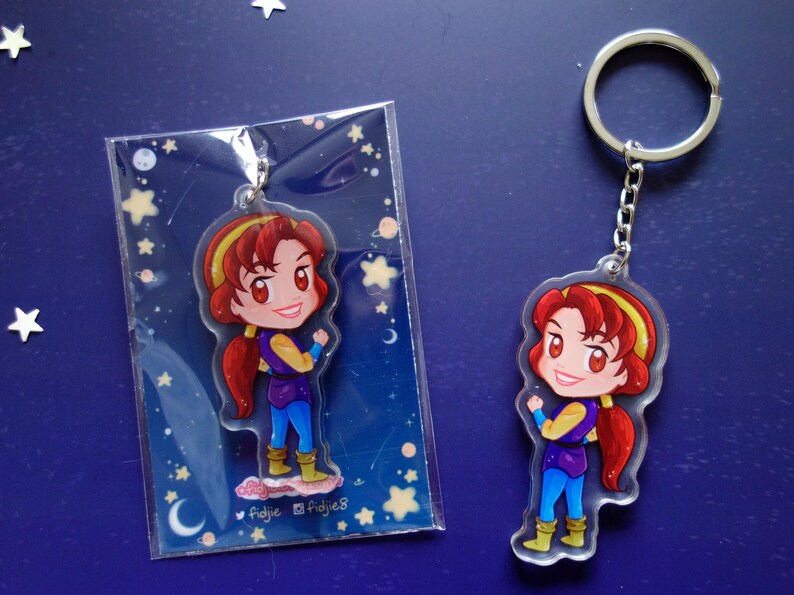 May include: A clear acrylic keychain featuring a cartoon character with red hair, a yellow headband, and a purple and blue outfit. The keychain is attached to a silver chain and ring. The character is also shown in a plastic sleeve.
