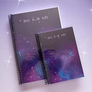 Puede incluir: Dos cuadernos de espiral con un estampado de galaxia en tonos morado y azul con estrellas blancas. El texto "SPACE IS THE PLACE" está impreso en blanco en las portadas. Los cuadernos están dispuestos en dos tamaños diferentes.