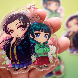 Hand holding a keychain with pink flower clasp of Jinshi and Maomao from manga series The Apothecary Diaries.