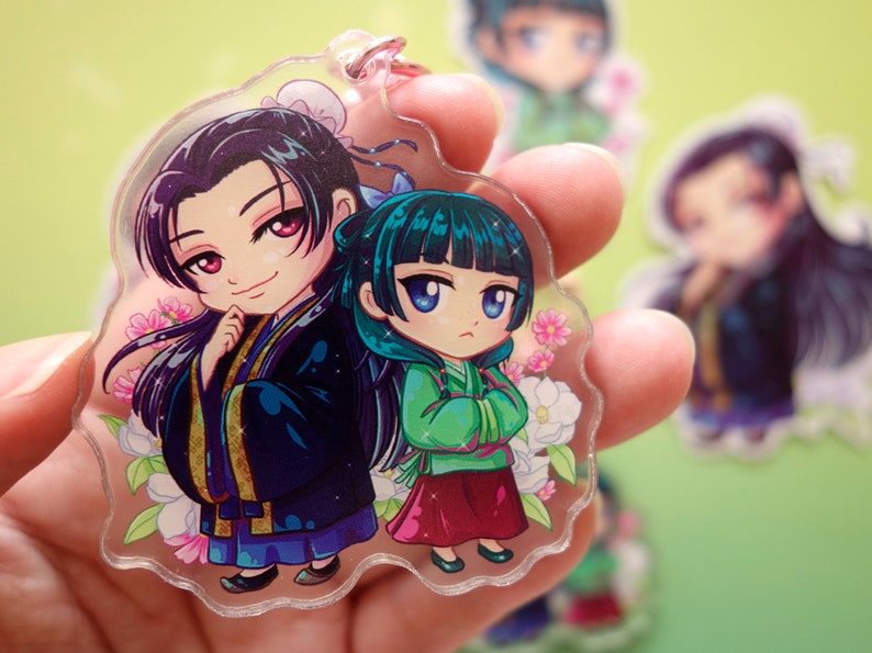 Hand holding a keychain with pink flower clasp of Jinshi and Maomao from manga series The Apothecary Diaries.