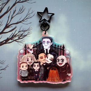 Puede incluir: Un llavero de acrílico colorido con los personajes de la familia Addams. Gómez, Morticia, Miércoles, Pugsley, el tío Fétido y la abuela están de pie frente a una puerta. El llavero tiene un amuleto negro en forma de estrella.