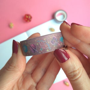 Close-up of a roll of washi tape with a pattern of Dungeons and Dragons elements on a purple background: dices, bow, axe, shield, book, potion, lute, etc