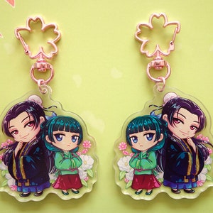 Clear acrylic keychains with pink flower clasp of Jinshi and Maomao from manga series The Apothecary Diaries, displayed both sides.