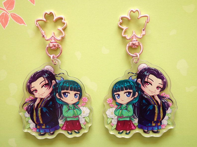 Clear acrylic keychains with pink flower clasp of Jinshi and Maomao from manga series The Apothecary Diaries, displayed both sides.
