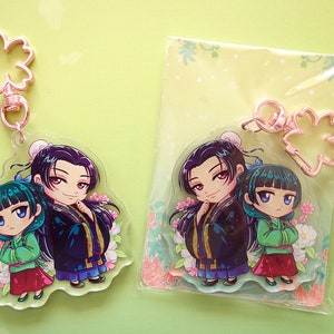 Clear acrylic keychains with pink flower clasp of Jinshi and Maomao from manga series The Apothecary Diaries. One is out of the envelope, and other is inside with a green back card.