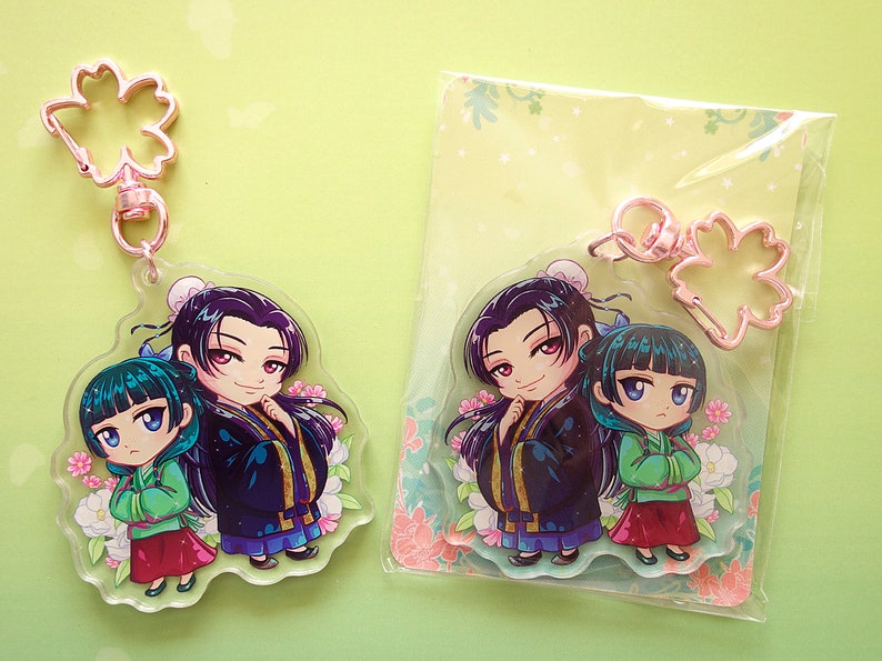 Clear acrylic keychains with pink flower clasp of Jinshi and Maomao from manga series The Apothecary Diaries. One is out of the envelope, and other is inside with a green back card.