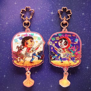 Könnte beinhalten: Zwei Schlüsselanhänger aus Acryl mit Figuren aus dem Animationsfilm Coco. Jeder Schlüsselanhänger hat einen rosa Rand, einen goldfarbenen oberen Clip und einen dekorativen Anhänger. Einer zeigt einen Jungen mit einem Hund, der andere eine Figur in einem roten Kapuzenpullover.
