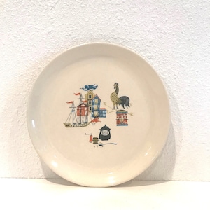 May include: A white ceramic plate with a colorful design featuring a rooster, a ship, houses, and a compass. The design is in shades of blue, red, yellow, and green.