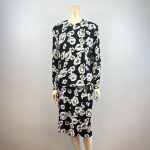 May include: A two-piece floral print outfit on a mannequin. The set includes a long-sleeved top and a knee-length skirt. The fabric is black with a pattern of large, white and gray flowers. The top has a button-up front.