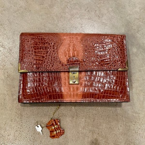May include: A brown leather clutch with a crocodile pattern and a brass lock and key. The clutch has a two-tone design with a lighter brown section in the center.