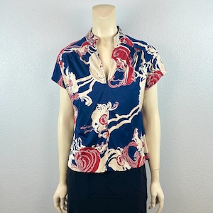 May include: A short-sleeved blouse with a mandarin collar. The blouse features a navy blue background with a repeating pattern of red and cream-colored floral and abstract designs. The blouse is displayed on a mannequin.