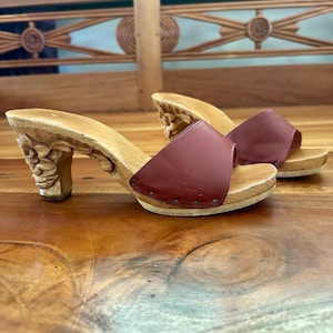 May include: A pair of slip-on clogs featuring carved light-colored wooden heels with floral detailing and a dark reddish-brown leather strap secured to the wooden base with small metal studs along the sides of the footbed.