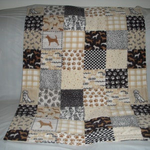 May include: A patchwork fleece blanket with a dog theme. The blanket is made up of various squares with different dog-related patterns, including paw prints, bones, and dogs. The blanket is beige, brown, and black.
