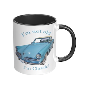 May include: White ceramic mug with a black rim and handle. The mug features a blue vintage car with a white roof and the text "I'm not old I'm Classic!"