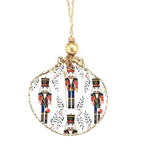 May include: A white, shell-shaped Christmas ornament featuring four toy soldier nutcracker figures in red, blue, and black uniforms. The ornament has a gold-colored trim and a natural fiber hanging cord with a gold bead.