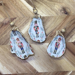 May include: Three oyster shell ornaments with gold edges and a nutcracker design. Each shell has a white background with a floral pattern and a string for hanging. The nutcrackers are in red, black, and blue uniforms.
