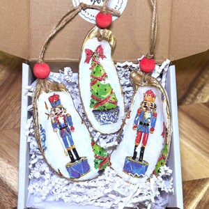 May include: Three Christmas ornaments made from oyster shells, each with a unique design. One features a green Christmas tree, and the other two depict a nutcracker soldier. The ornaments are strung with twine and red beads, presented in a box.