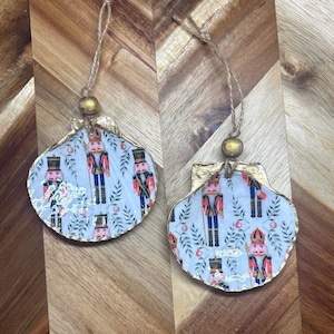 May include: Two seashell Christmas ornaments. Each ornament features a printed design of a nutcracker soldier on a light blue background with gold accents. The shells are trimmed with gold paint and have a jute string for hanging.