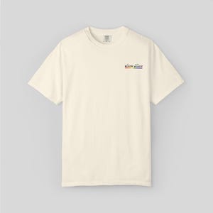 May include: Cream-colored short-sleeve t-shirt with the text "Same Team" in a rainbow color scheme. The shirt has a crew neck and a small tag at the neckline.