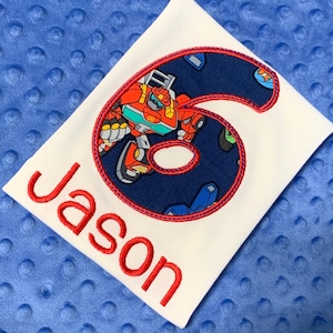 May include: White t-shirt with a red embroidered number six. The number six is filled with a blue fabric featuring a cartoon robot. The name "Jason" is embroidered in red below the number.