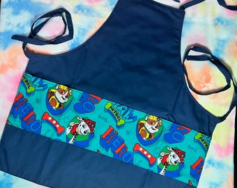 May include: A blue child's apron with a pocket and adjustable straps. The pocket has a green fabric with a repeating pattern of cartoon dogs from the Paw Patrol cartoon. The fabric says "PAW PATROL" and "HERO" in a white font.