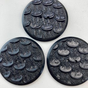 Op de afbeelding: Drie zwarte ronde knoppen met een getextureerd oppervlak en een patroon van kleine, verhoogde, halfronde vormen. Elke knop heeft twee gaten in het midden.