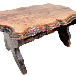 May include: A wooden footstool with a carved top and turned legs. The stool is dark brown and has a rustic appearance.