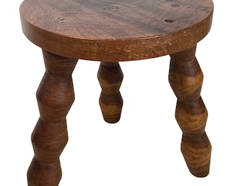 May include: A wooden stool with a round top and three legs. The legs are turned and have a bulbous shape. The stool is made of dark brown wood.