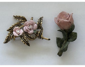 Puede incluir: Dos broches de rosas. Uno es de metal dorado con dos rosas rosas y detalles verdes. El otro es una sola rosa rosa con hojas y tallo verdes. Ambas son joyas con temática floral.