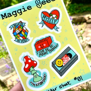 May include: A sticker sheet with six colorful stickers. The stickers feature a red heart with the word "MYSELF" on it, a red brick with a smiley face and the text "I CAN BE SO MANY THINGS!" a mushroom with a smiley face and the text "I'M A FUN DUDE", a cassette tape with a smiley face and the text "PLEASE BE KIND TO YOUR MIND!" a Rubik's cube with a smiley face and the text "I BELIEVE IN YOU!" and a blue and green Rubik's cube with the text "Maggie Seee" on the top. The sticker sheet is labeled "Sticker Sheet #01".