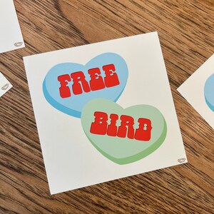 May include: A blue and green heart-shaped candy with the words "FREE BIRD" in red letters.
