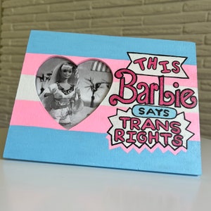 May include: A wooden picture frame painted in pink, blue, and white with a heart-shaped cutout. The cutout features a black and white photo of a Barbie doll. The frame has the text "This Barbie Says Trans Rights" in pink and white.