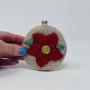 May include: A white beaded clutch with a red flower design. The flower has green leaves and a yellow center.
