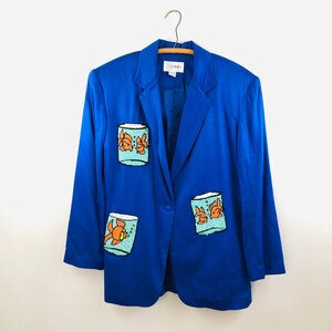 May include: A royal blue blazer with three embroidered patches of goldfish in bowls. The blazer has a single button closure.