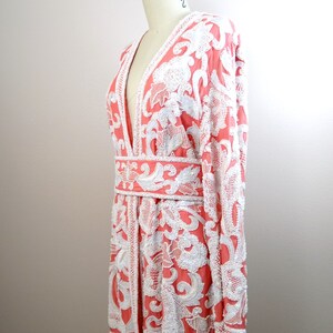 Op de afbeelding: Een koraalroze en witte pareljas met een V-hals en lange mouwen. De jas heeft een bloemenpatroon en een riem in de taille.