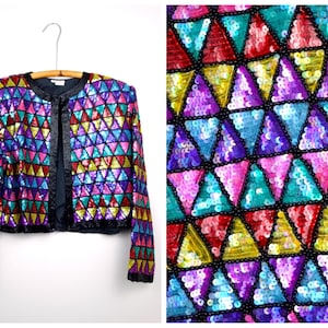 Pode incluir: Uma jaqueta colorida de lantejoulas com um padrão geométrico de triângulos em tons de rosa, roxo, azul, verde, vermelho e amarelo. A jaqueta tem um forro preto e mangas compridas.