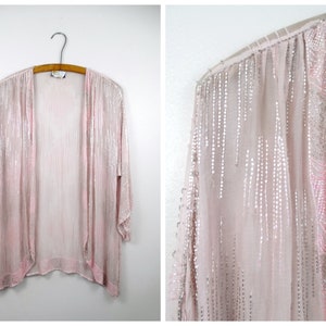 Puede incluir: Un cárdigan de color rosa claro con flecos largos y cuentas de color plateado. El cárdigan es de color rosa claro y está decorado con cuentas de color plateado.