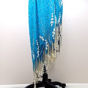 May include: A turquoise sequined dress with a silver fringe and white bead accents. The dress is on a black stand with wheels.