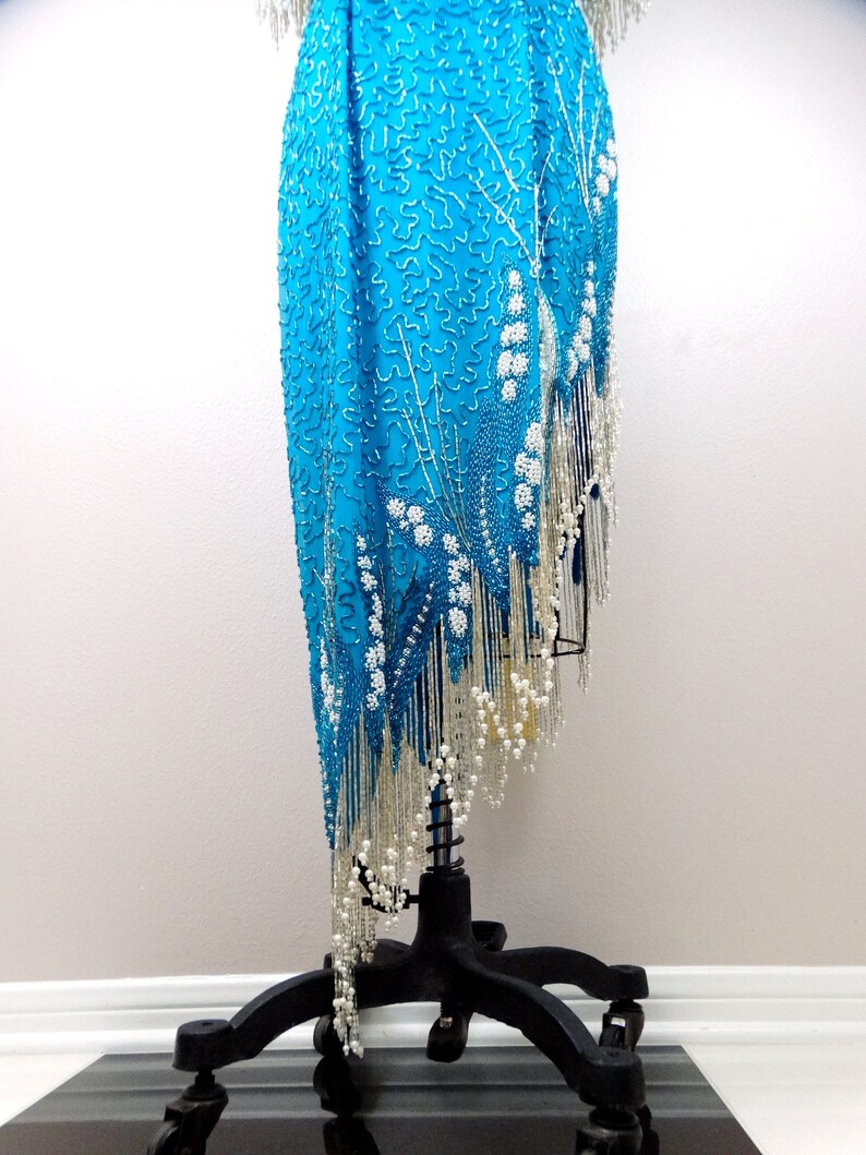 May include: A turquoise sequined dress with a silver fringe and white bead accents. The dress is on a black stand with wheels.