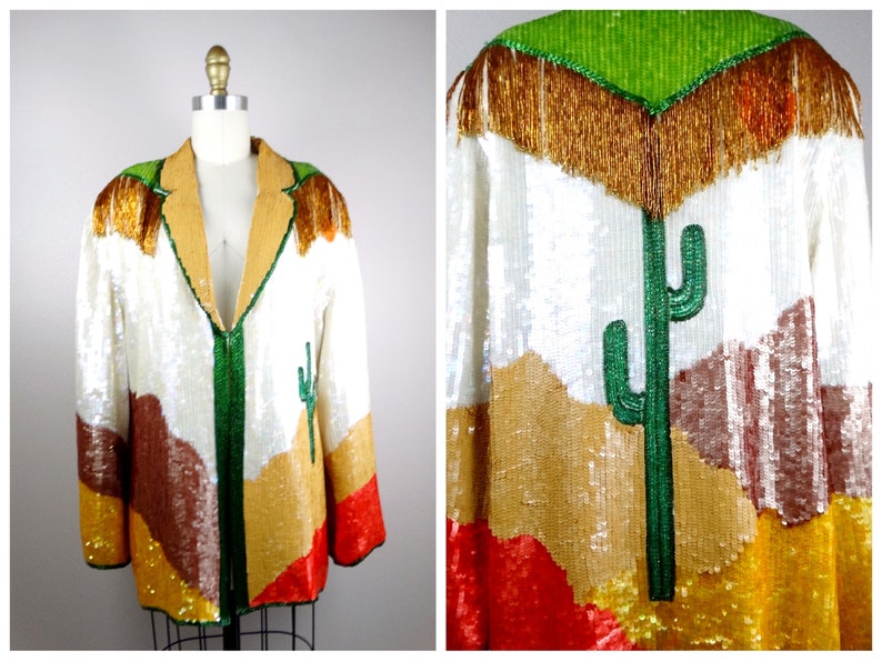 May include: A white sequined jacket with a desert landscape design. The jacket features a green cactus, brown mountains, and gold and red accents. The jacket has a green zipper and a gold fringe collar.