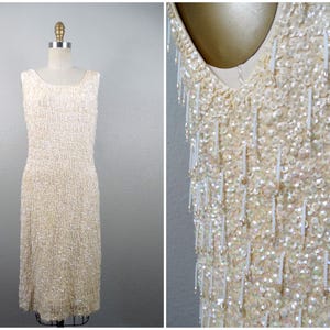 Può includere: Un abito bianco senza maniche realizzato con paillettes e perline. L'abito ha un dettaglio di frange di perline.