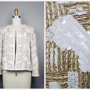 May include: A white and gold sequined jacket with a geometric pattern. The jacket is cropped and has long sleeves. The sequins are arranged in a pattern of squares and rectangles.