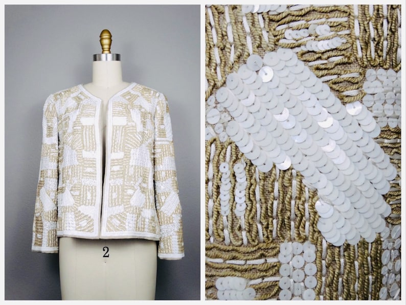 May include: A white and gold sequined jacket with a geometric pattern. The jacket is cropped and has long sleeves. The sequins are arranged in a pattern of squares and rectangles.