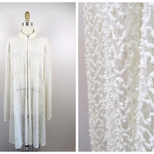 Puede incluir: Una chaqueta blanca de encaje con un diseño de cuentas de perlas. La chaqueta tiene una silueta larga y fluida y un cuello de pie.