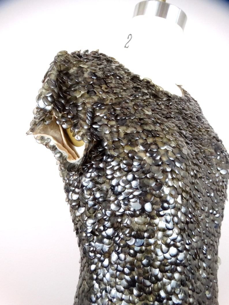 May include: A close-up of a garment made of overlapping, iridescent, grey, plastic scales. The garment is on a dress form.