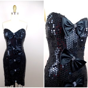 May include: A black strapless sequined dress with a fitted silhouette. The dress features multiple black satin bows with rhinestone accents.