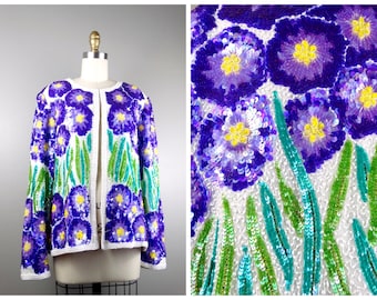 Op de afbeelding: Een witte jas zonder sluiting versierd met pailletten, met een bloemenmotief. De jas toont grote paarse bloemen met gele centra en groene en turquoise bladeren. Het kledingstuk wordt op een mannequin gepresenteerd.