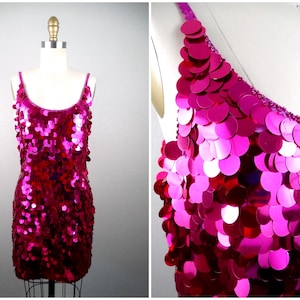 May include: A vibrant fuchsia sequin dress with thin straps. The dress is covered in large, round sequins that catch the light, creating a shimmering effect. The dress is displayed on a mannequin, showcasing its form-fitting design.