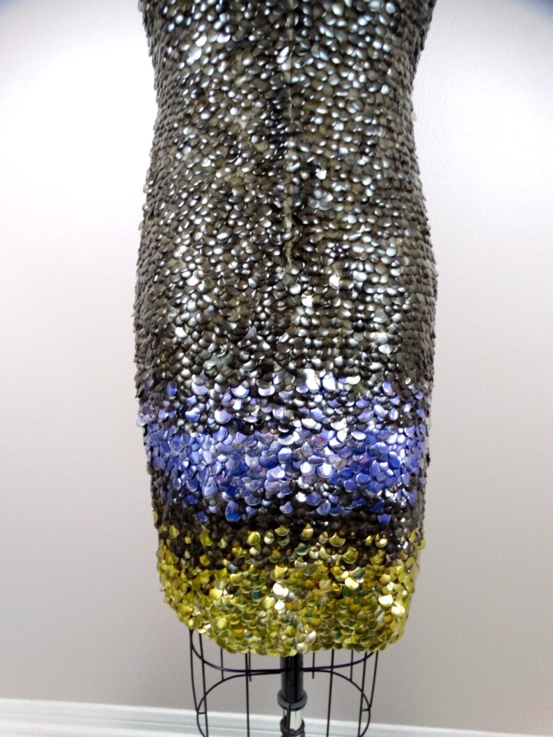 May include: A sequined dress with a silver, purple and gold colour scheme. The dress is made of small, overlapping sequins that create a shimmering effect.